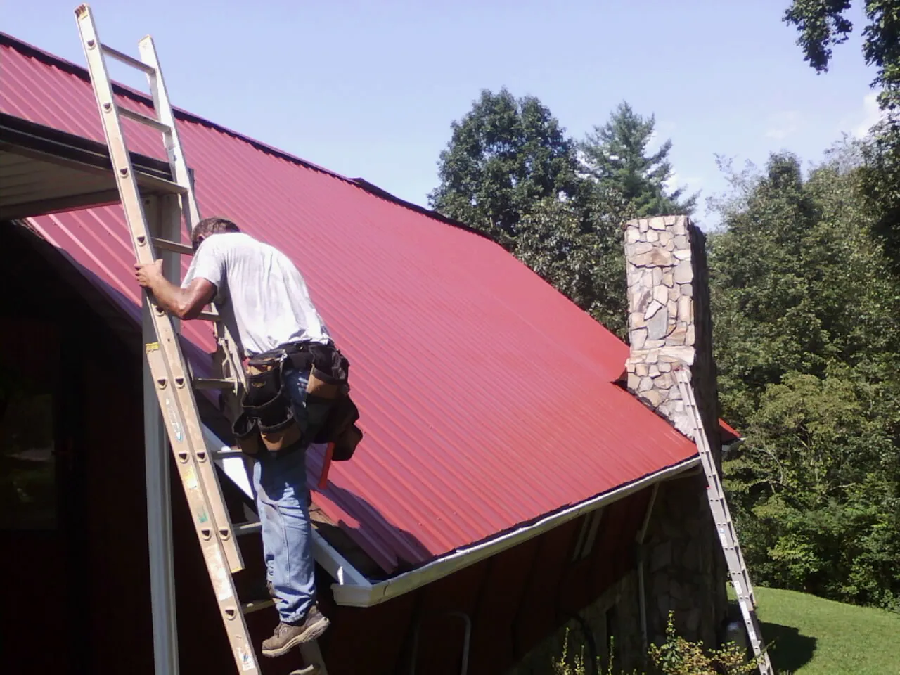 Roofer on a ladder beside a red metal roof and stone chimney — Soffit & Fascia Repair by Roofing Brothers in Santa